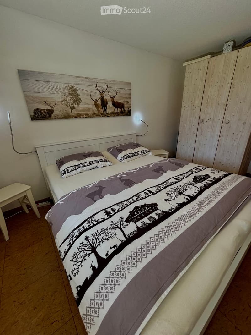 Bedroom with a double bed, pillows, bedspread, a small stool, a wooden wardrobe, and a large picture on the wall.