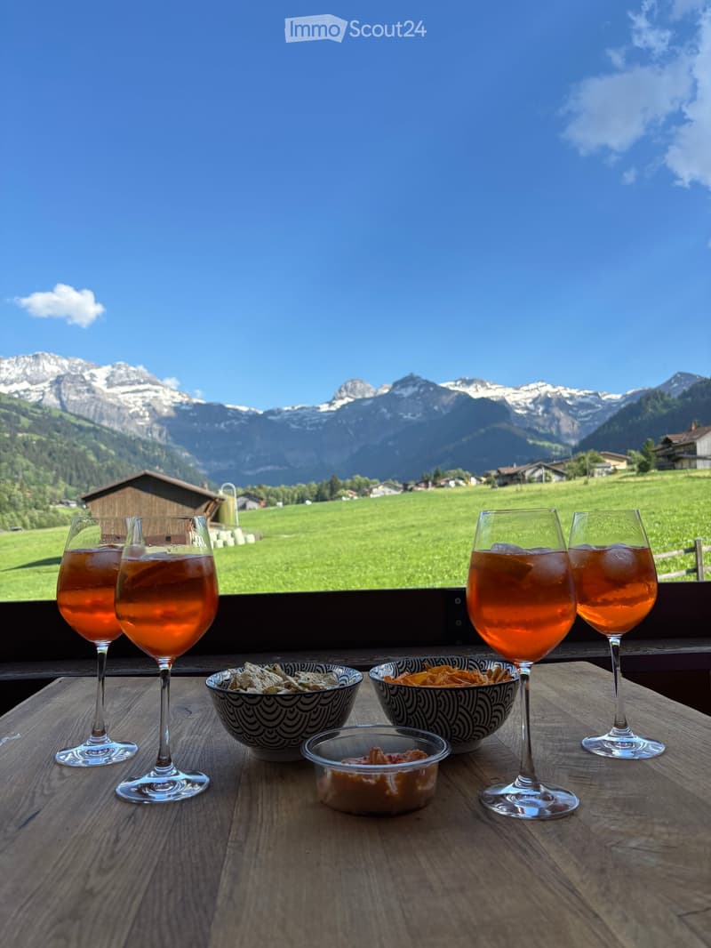 Outdoor table with wine glasses and bowls, overlooking a scenic mountain landscape with green fields and houses.