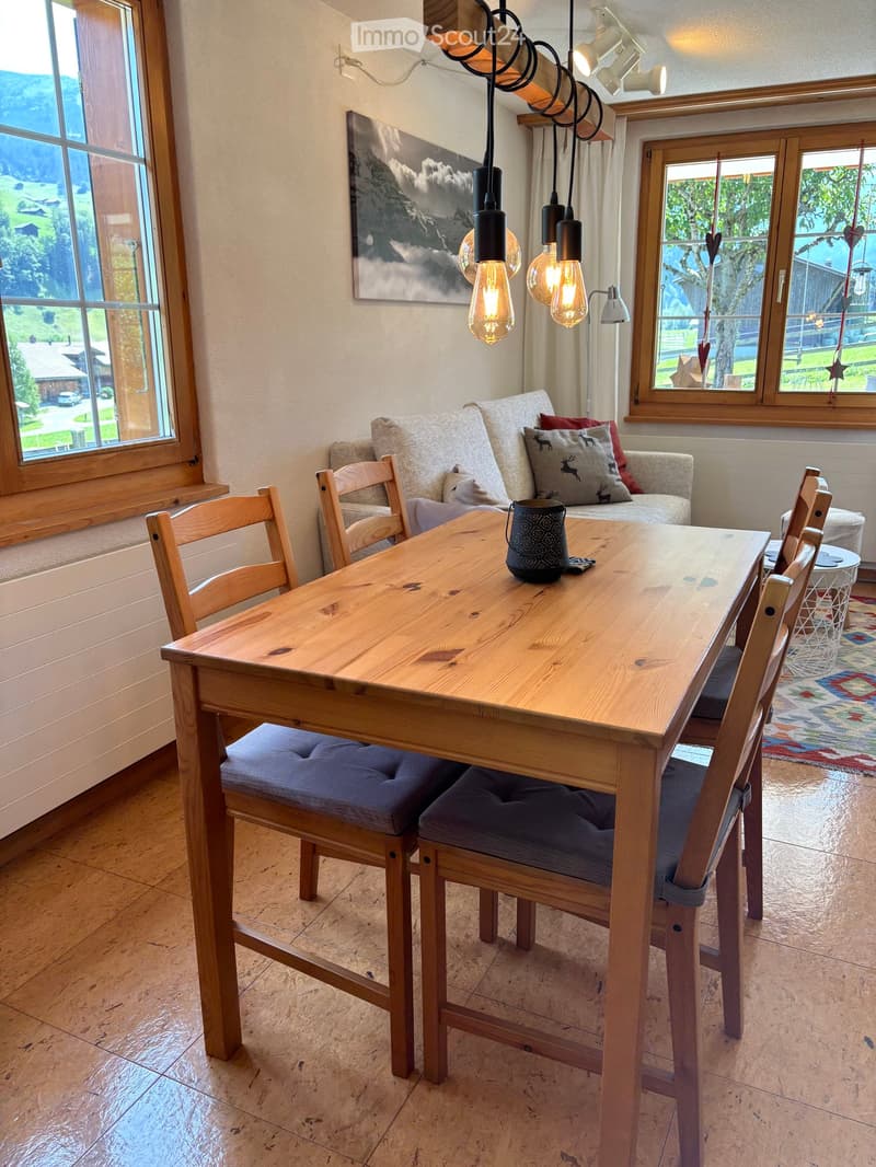 Bright dining area with wooden table, cushioned chairs, sofa, pendant lights, and scenic windows overlooking a landscape.