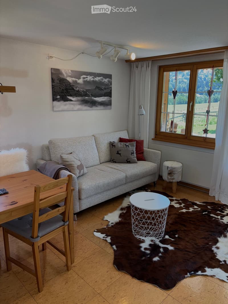 A cozy living room with a grey sofa, wooden table, chair, and a cowhide rug. Large windows offer a scenic view, complemented by modern lighting and decorative elements.