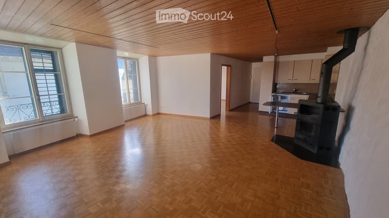 large empty room with wooden floors, fireplace on the right, large windows with balconies, open kitchen