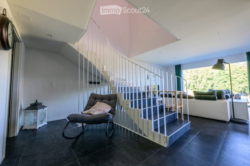 Modern living room with a staircase, dark tiled floor, white walls, and large windows. Features include a white sofa, a rocking chair with a cushion, a small table, a floor lamp, and a decorative lantern.