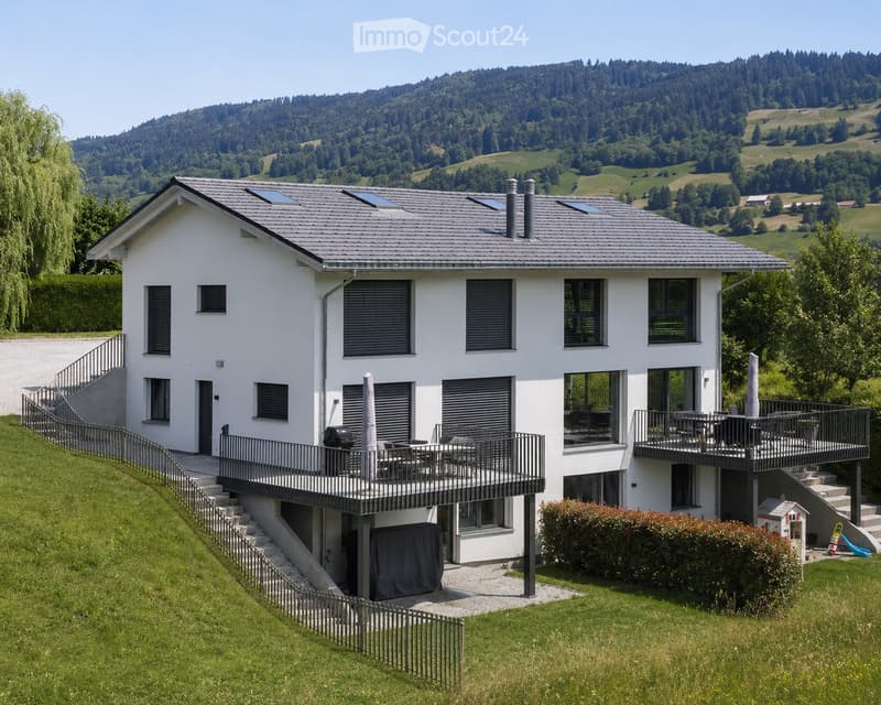 Two-story white house with grey roof, multiple windows, balconies, and outdoor seating areas. Surrounded by green lawn and trees with a scenic mountain backdrop.
