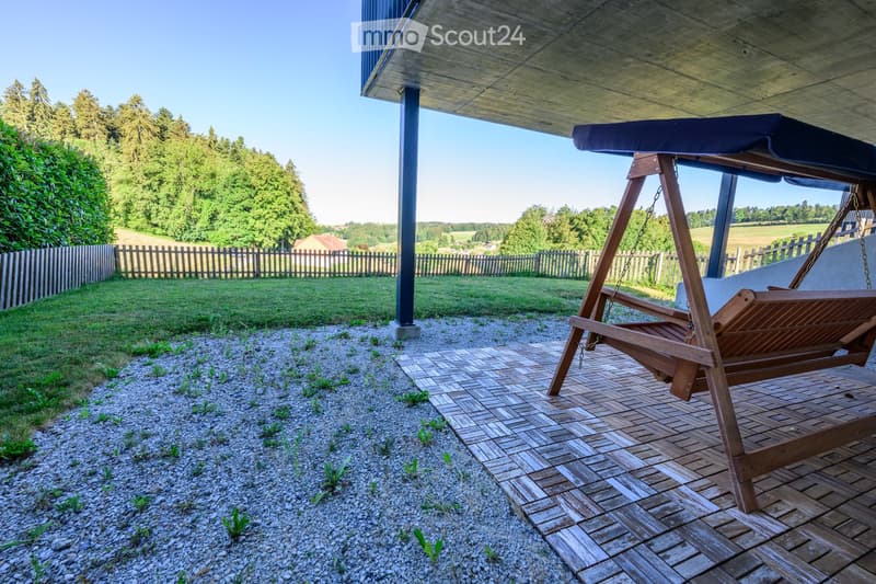 A wooden swing chair sits on a wooden deck under a covered outdoor area, surrounded by a gravel area and a grassy lawn with trees in the background.