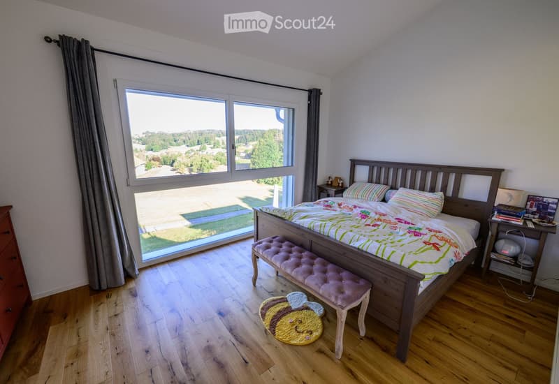 Bright bedroom with wooden flooring, large window offering a scenic view, a wooden bed with colorful bedding, a matching bench, a nightstand, and a decorative rug.