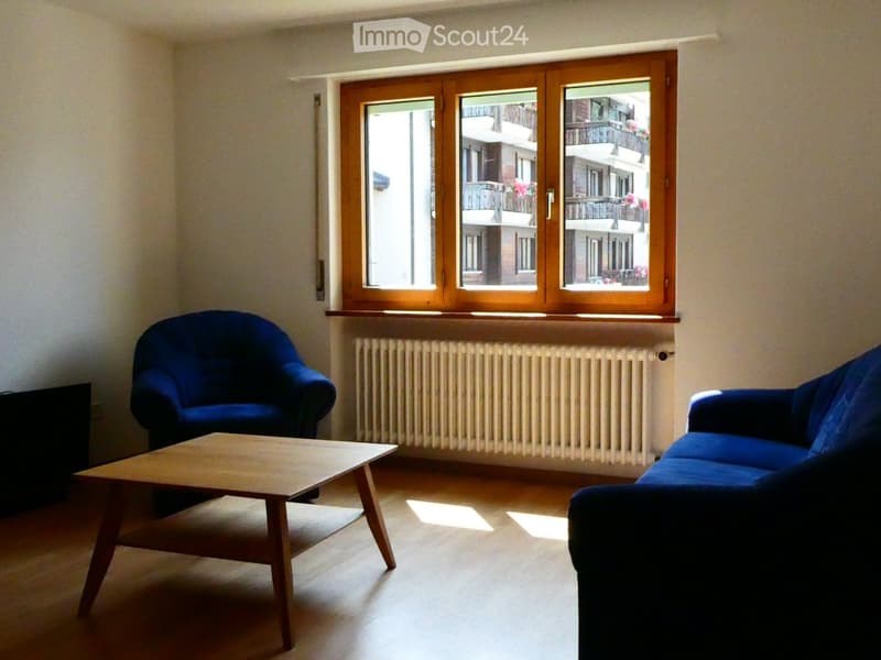 Living room with wooden floor, large window with wooden frame, radiator, blue sofa, armchair, and coffee table.