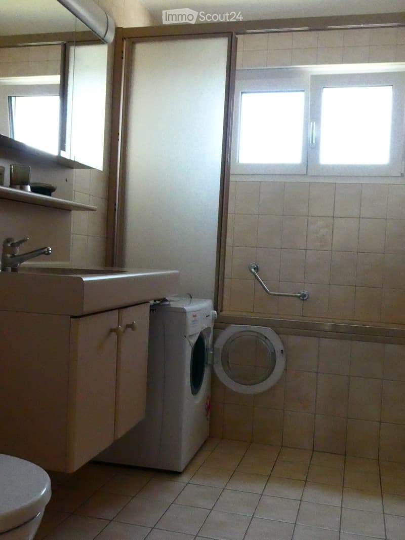 Bathroom with a sink, faucet, cabinet, washing machine, and toilet. The room has tiled walls and floor, with a window providing natural light.