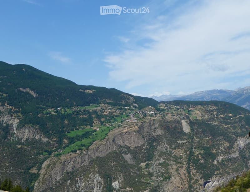 A panoramic view of a mountainous valley with scattered houses, green fields, and rocky terrain under a clear blue sky.