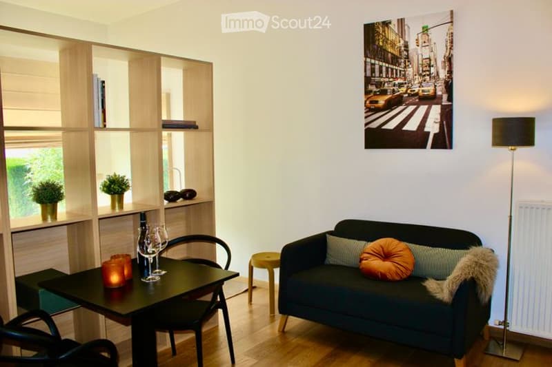A modern, well-lit living room with a dark blue sofa, a small black dining table with chairs, a floor lamp, and built-in wooden shelves holding potted plants and decorative items.