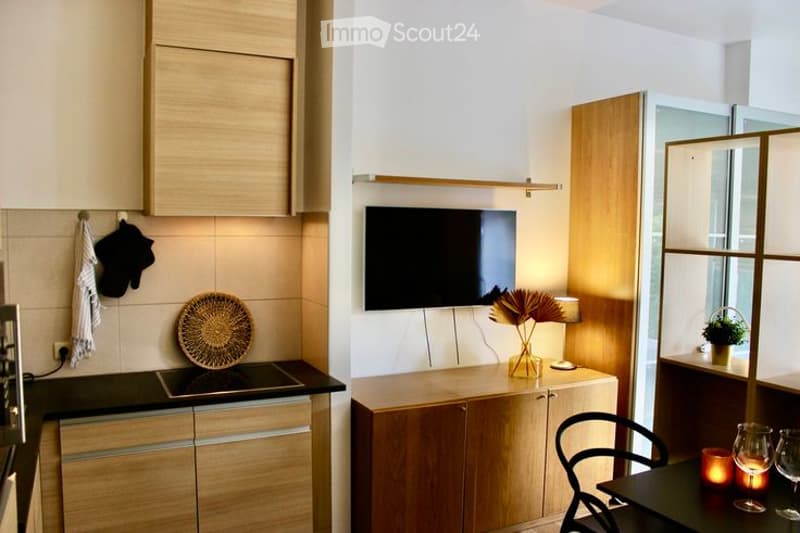 Modern kitchen and living area with light wood cabinets, induction stove, wall-mounted TV, small dining table with chairs, decorative vase, candle, and wine glasses.