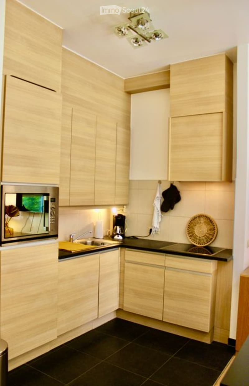 Modern kitchen with light wood cabinets, black countertops, and stainless steel appliances including a microwave and coffee maker. Features a sink with a faucet, a built-in oven, and a decorative woven basket on the counter.