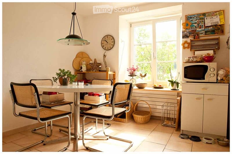 Bright kitchen with a dining area, white cabinets, a microwave, and a table with chairs. Natural light streams in from large windows, and decorative items like potted plants and a clock adorn the space.