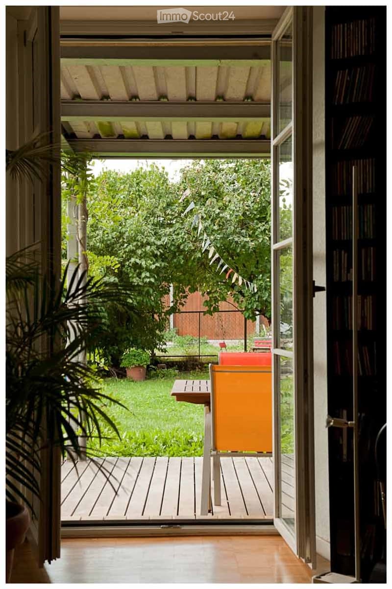 Open balcony with wooden decking, orange chair, small table, lush greenery visible through large glass doors.
