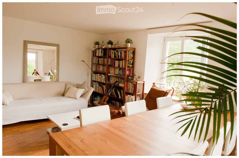 Bright living room with white sofa, wooden dining table, bookshelf, and large windows letting in natural light.