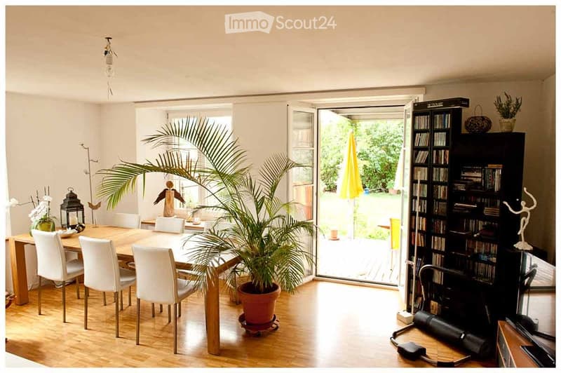 Bright, open dining area with a large potted plant, a wooden dining table with white chairs, a tall bookcase, and a view to a terrace.