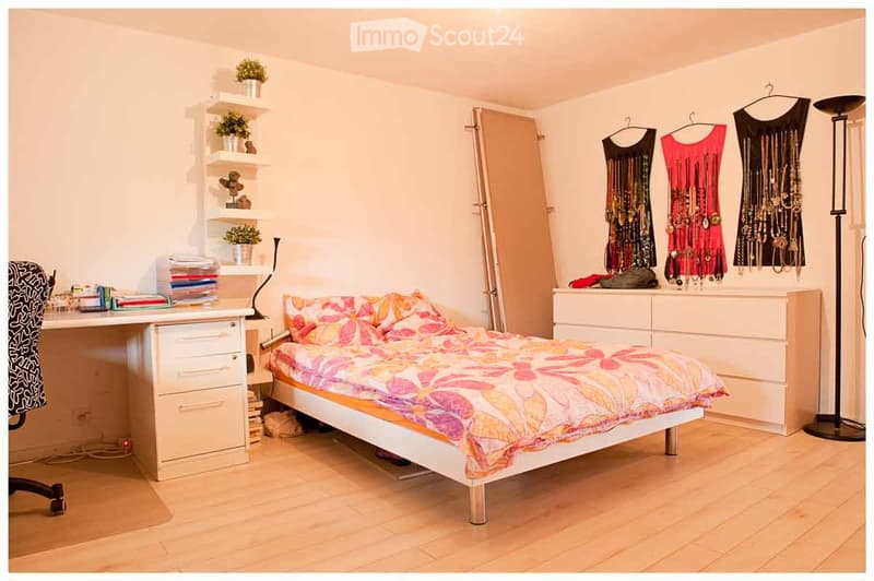 Bright bedroom with a white bed, colorful floral bedding, a white desk with a chair, shelves with plants and books, a dresser with a mirror, and a jewelry rack with necklaces and dresses.
