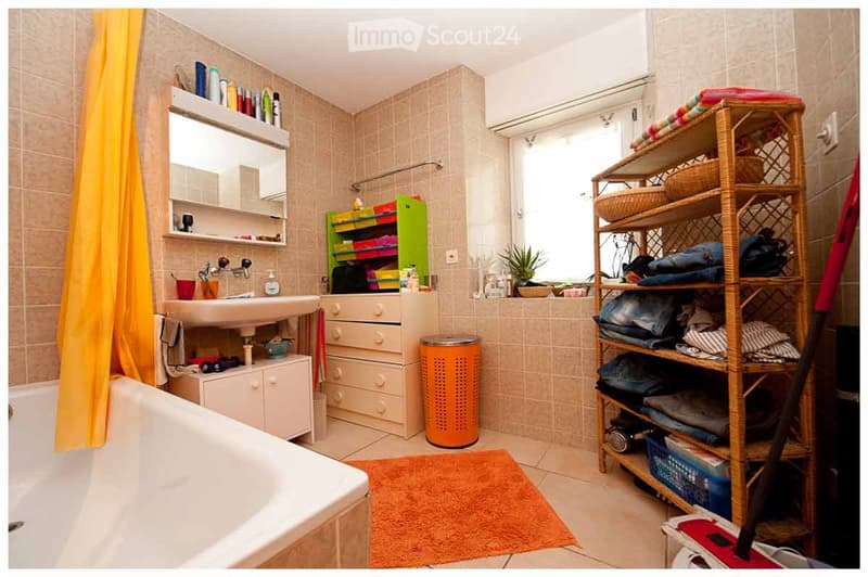Bathroom with a bathtub, sink, mirror, cabinet, wicker shelf, trash bin, and a rug. The room features tiled walls and a window with a view.