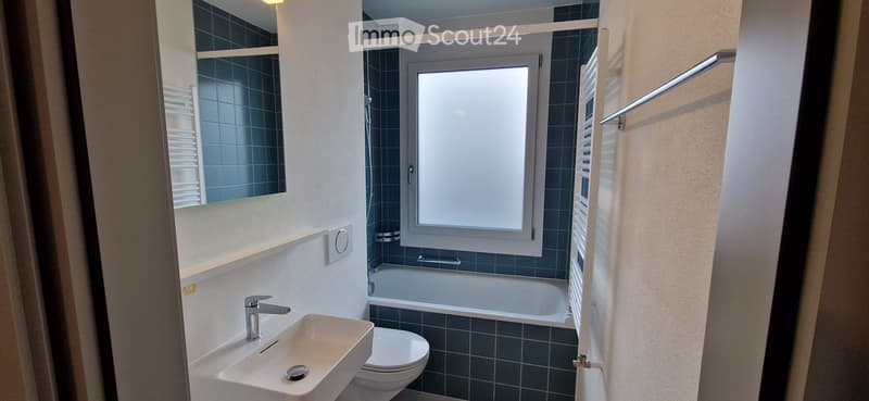Modern bathroom with a white sink, chrome faucet, toilet, bathtub, and a large window. Blue tiled walls and a radiator are visible.