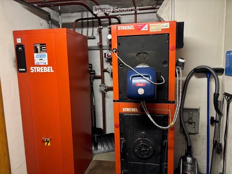 Utility room with two orange Strebel boilers, a vacuum cleaner, and exposed pipes.