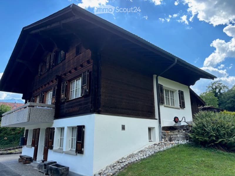 Traditional Swiss wooden house with white lower facade, multiple windows with shutters, and a balcony on the left side.