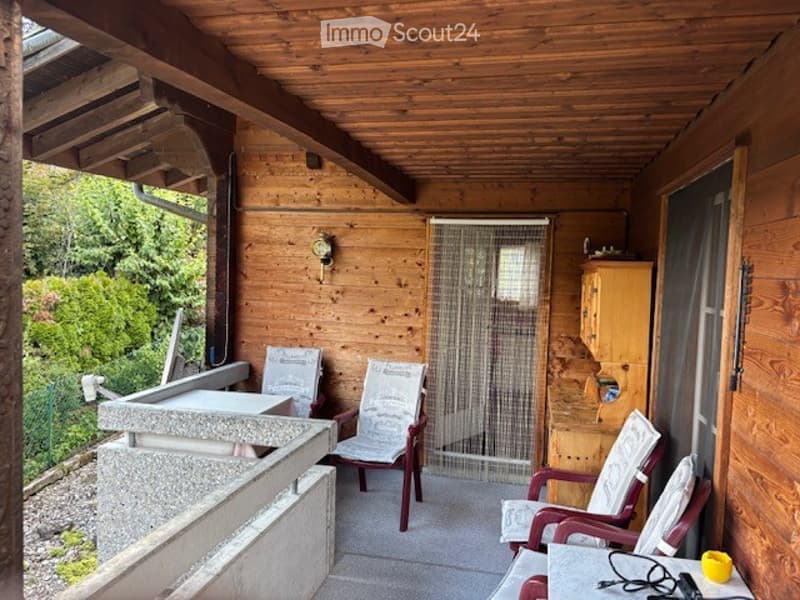 Wooden balcony with a wooden ceiling, wooden walls, and a small table with chairs. There is a small table and a few chairs, and a small cabinet on the right side.
