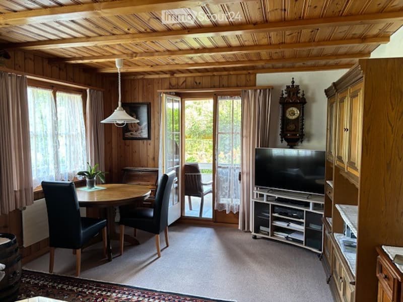 A cozy, wooden-paneled room with a round wooden dining table and four black chairs, a television on a stand, a large wooden cabinet, a wall-mounted clock, a pendant light, and a small plant on the table. Large windows with curtains lead to a balcony or ou