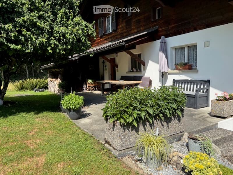 A well-maintained garden area in front of a house, featuring a wooden bench, a table with chairs, potted plants, and a stone pathway.