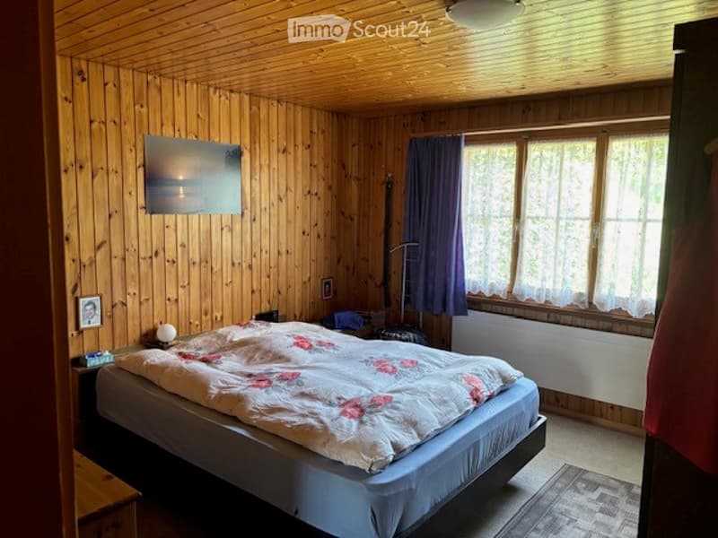 Cozy wooden-paneled bedroom with a large bed, nightstand, lamp, picture frame, radiator, and curtains on the windows.