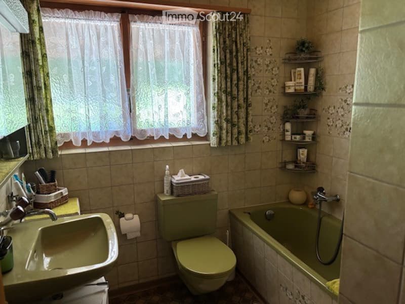 Bathroom with green tiles, a window with white lace curtains, a green sink, a green toilet, a green bathtub, and shelves with toiletries.