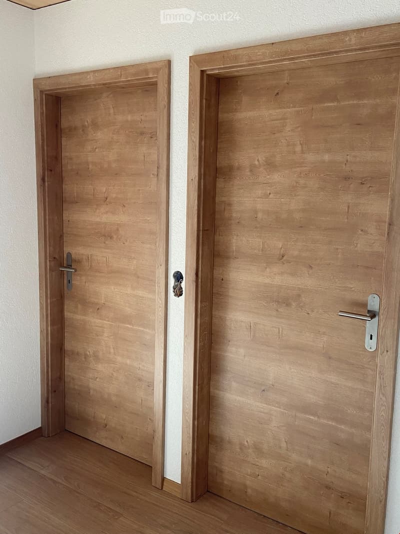 A narrow hallway with two wooden doors, one slightly ajar, featuring a light-colored wooden floor and white walls.