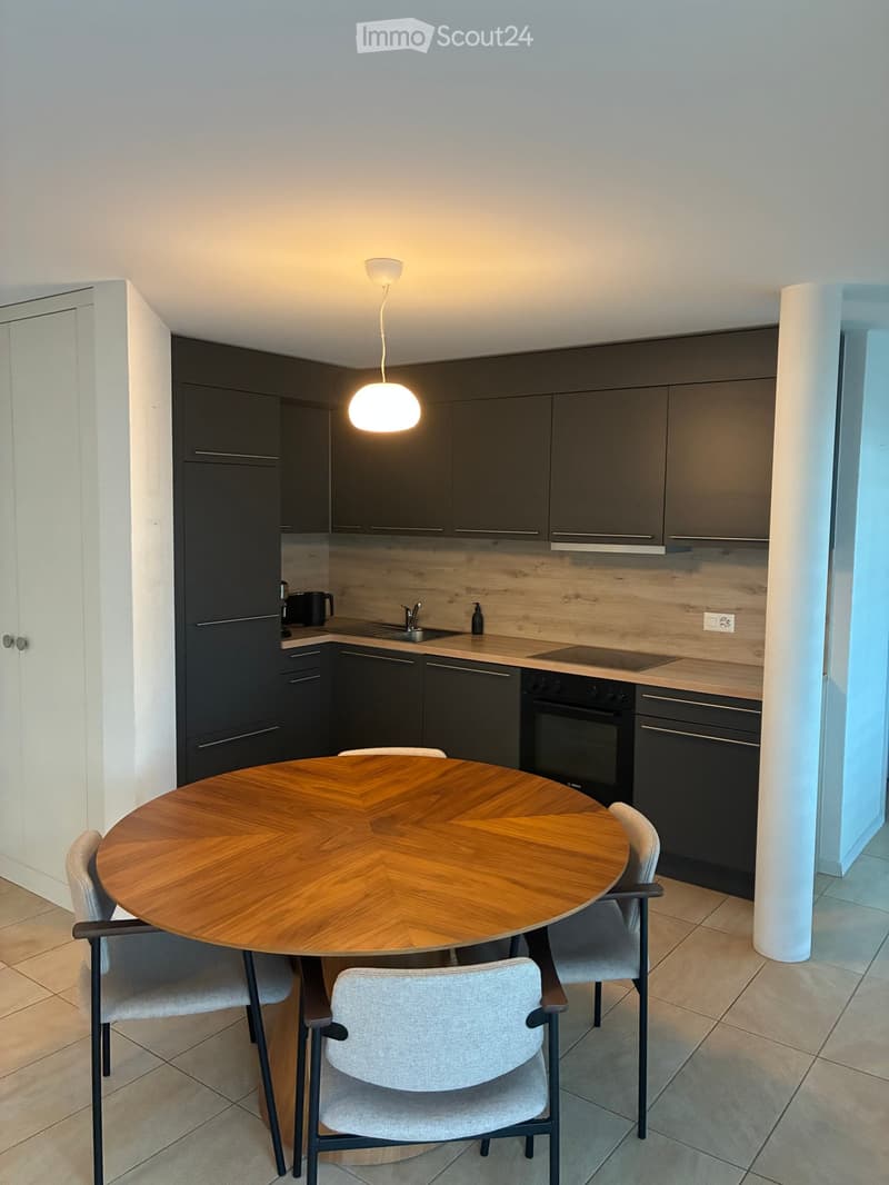 Modern kitchen with dark grey cabinets, a round wooden dining table with four chairs, and built-in appliances including an oven and dishwasher.