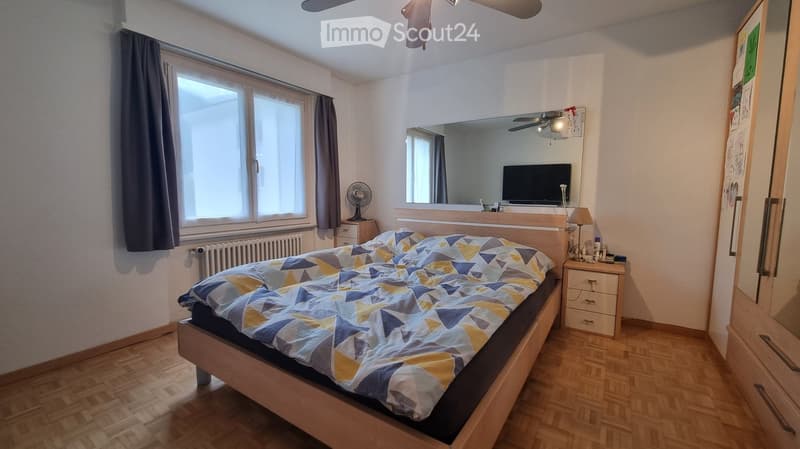 Well-lit bedroom with wooden flooring, large window with curtains, bed with geometric-patterned quilt, mirror, television, fan, and nightstand with lamp.