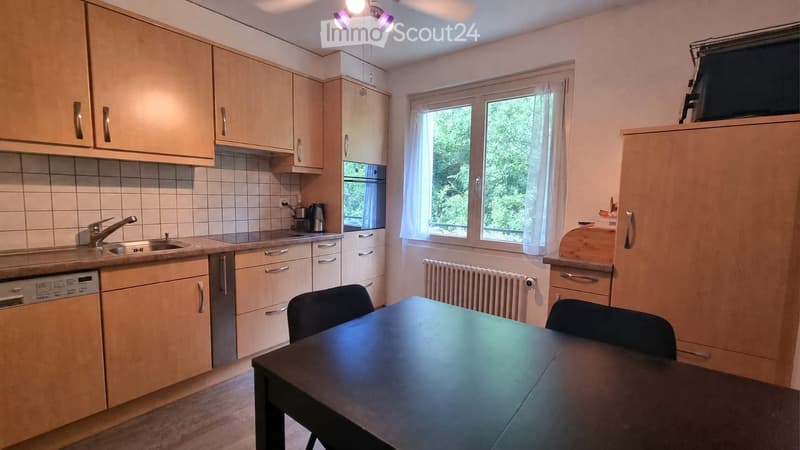 Bright kitchen with light wooden cabinets, a sink, dishwasher, and a dining table with chairs. Large windows provide natural light.