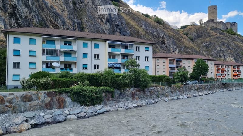 Multi-story apartment building with balconies and blue shutters, situated near a river with rocky banks and a castle on a hill in the background.