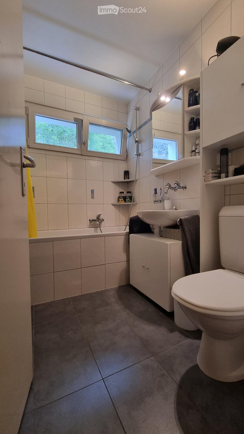 Modern bathroom with a bathtub, toilet, sink, and built-in cabinet. Features tiled walls, dark grey floor tiles, and a window with blinds.