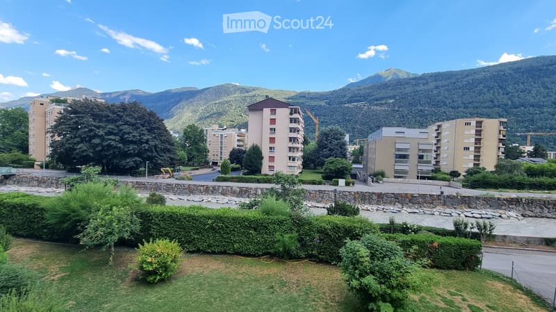 View of a residential area with multiple apartment buildings, surrounded by greenery and mountains in the background.