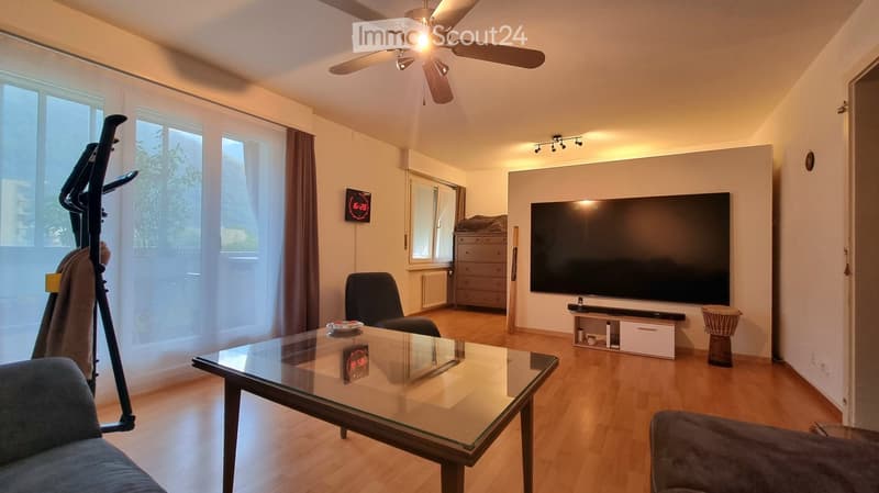 Modern living room with wooden flooring, large windows, a ceiling fan, a flat-screen TV, a glass coffee table, a sofa, a chair, a cabinet, and a fitness bike.
