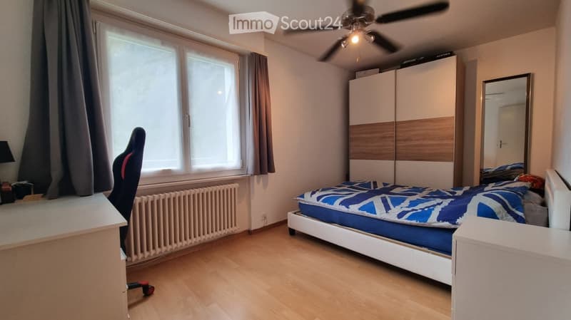 Bright bedroom with wooden flooring, large window with curtains, bed with blue and white bedding, wardrobe, chair, and a ceiling fan with light.