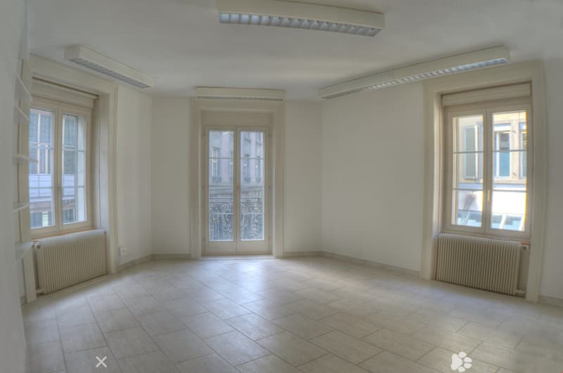 This is a room with white walls, tiled flooring, and large windows with double glazing and white frames. There are two white radiators below the windows and ceiling-mounted LED lights. The room appears to be empty with no furniture or decor visible.