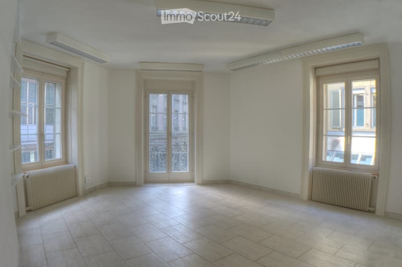 This is a room with white walls, tiled flooring, and large windows with double glazing and white frames. There are two white radiators below the windows and ceiling-mounted LED lights. The room appears to be empty with no furniture or decor visible.