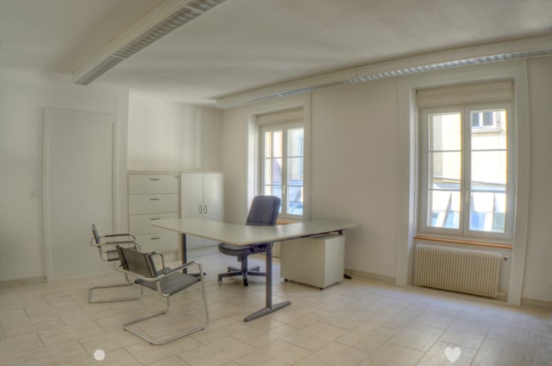 a table, two gray office chairs, a gray office chair, white wooden cabinets, two windows, and a white radiator