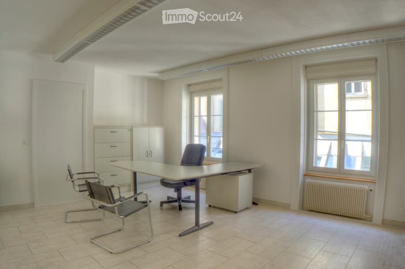 a table, two gray office chairs, a gray office chair, white wooden cabinets, two windows, and a white radiator
