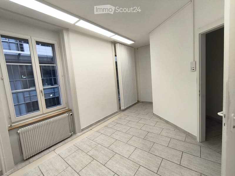 Empty room with tiled floor, white walls, large windows, radiator, and a closed door leading to another space.