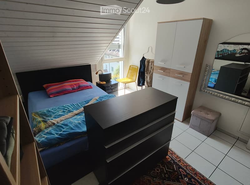 Bedroom with a bed, nightstand, chair, wardrobe, mirror, and a small stool. The room features a slanted ceiling and a window with a view.