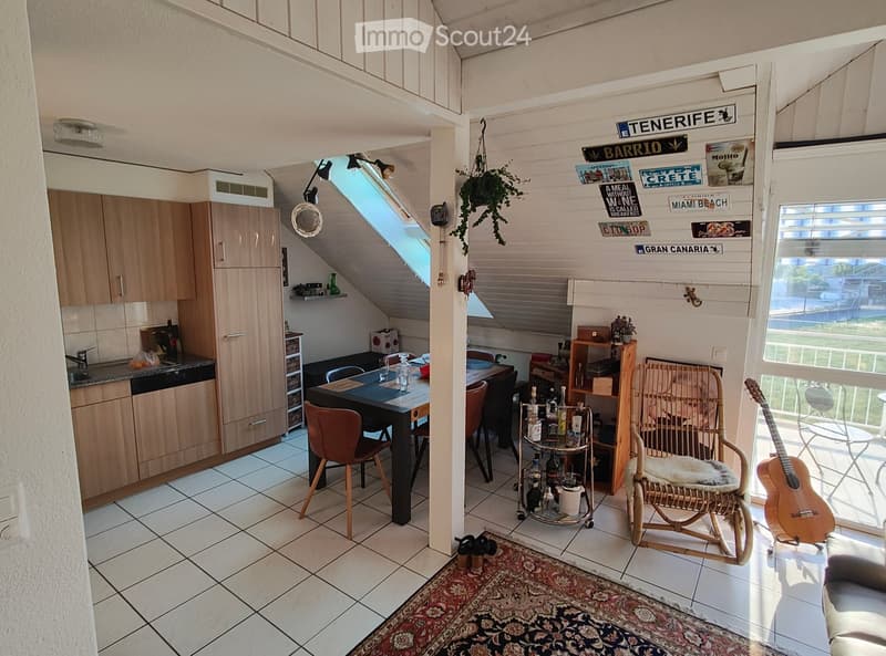 The image shows a cozy, well-furnished room with a kitchen area, dining table, and seating area. The room features wooden cabinets, a sink, a dining table with chairs, a wicker chair, a guitar, and a small cart with bottles. The space is decorated with a