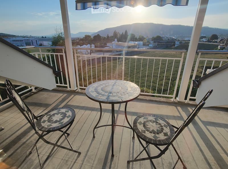 Balcony with a round mosaic table and two matching mosaic chairs, overlooking a scenic landscape with hills and a cityscape.