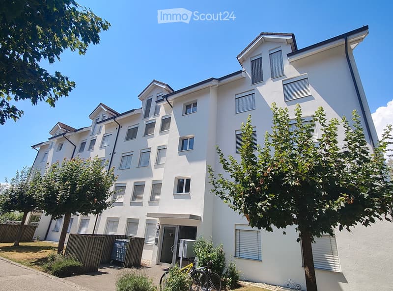 Modern white multi-story residential building with multiple windows, balconies, and surrounding greenery including trees and shrubs.