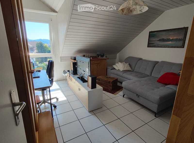 A cozy attic room with a slanted wooden ceiling, tiled floor, and large window offering a scenic view. The room features a grey sectional sofa with cushions, a wooden desk with a computer setup, and a hanging lamp providing ambient lighting.
