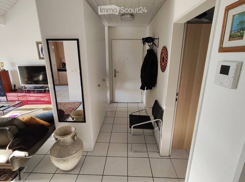 Hallway with tiled floor, white walls, a large mirror, a coat rack, a vase, a chair, and a television visible in the adjacent room.