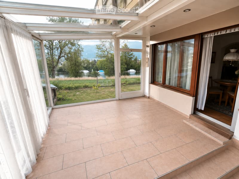 Bright, tiled balcony with large windows and sliding doors, offering a scenic view of a lake and surrounding greenery.